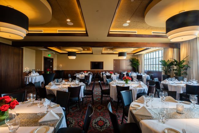 Truluck's Rosemont Stone Crab Room view from the the far end of the room looking across to the far wall where a TV is on the wall. Tables are dressed in white linen with silver table runners, candles and flowers.