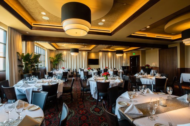 Truluck's Rosemont Stone Crab Room view from the main entry side across the room to the far wall where a TV is on the wall. Tables are dressed in white linen with silver table runners, candles and flowers.