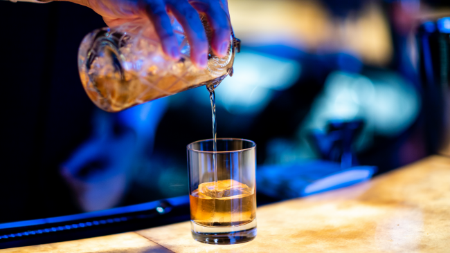 Bartender hand pouring bourbon over an ice block in a rocks glass from a glass shaker tin with strainer