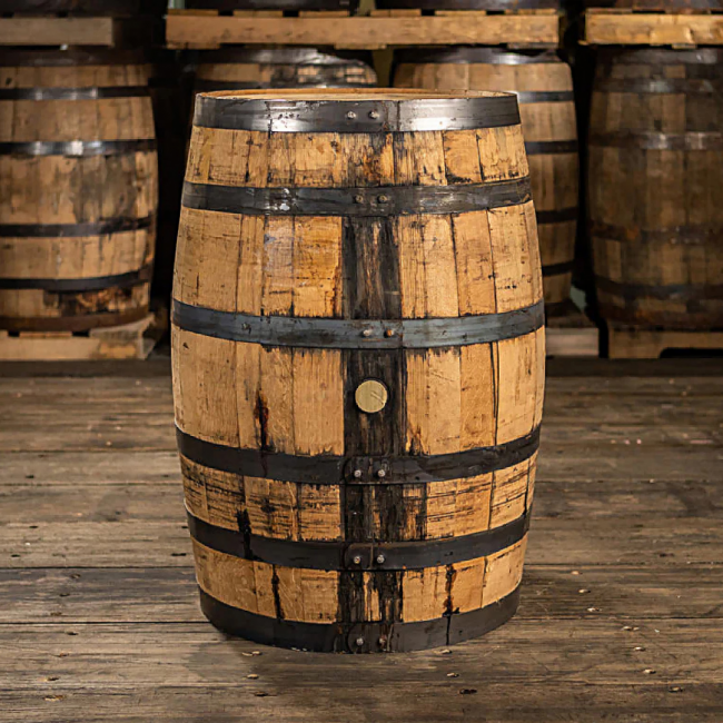 A wooden bourbon barrel in a store room with a wood floor and several more wooden barrels in the background.