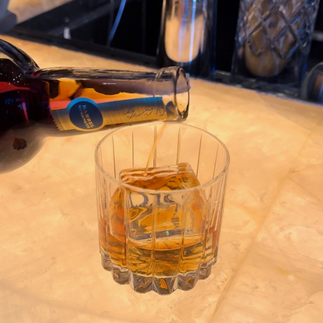 A bottle of Truluck's reserve whiskey being poured into a rocks glass with a square ice block in the glass.