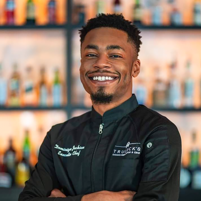 Chef Dominque of Truluck's Arboretum in a black chef coat with his arms crossed standing in front of the bar