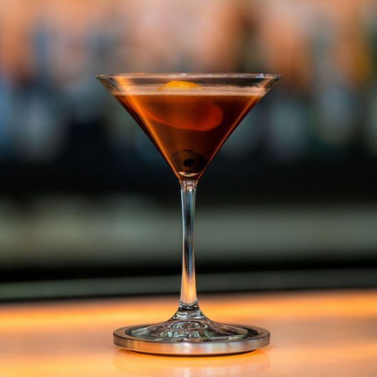 Truluck's reserve Manhattan cocktail in a martini style glass sitting on a silver coaster. the drink is garnished with a cherry and orange twist