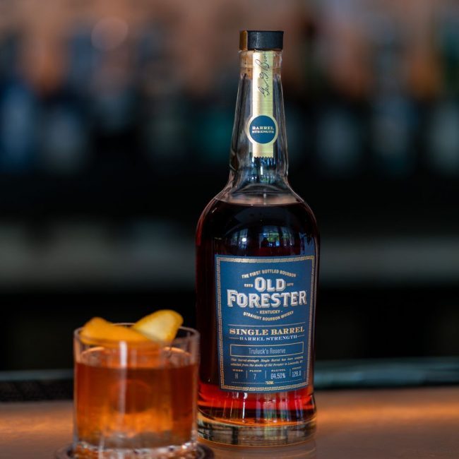 Old Forester Kentucky straight bourbon whiskey. Single barrel strength Truluck's Reserve 129 proof bottle with a navy and gold labeled and an old fashioned cocktail in a rocks glass sitting on the bartop next to the bottle.