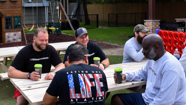 A group of veterans sitting at a picnic table at the PTSD Foundation of America's Camp Hope facility. They are enjoying Katz coffee in logo coffee cups. There is a playground in the background of the yard.