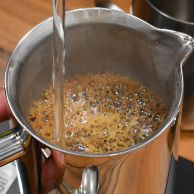 An overhead shot of hot water being poured over coffee grounds into a stainless steel French press pitcher.