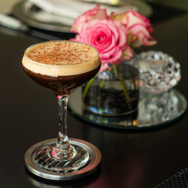 An espresso martini sitting on a silver coaster on a table with fresh pink roses in the background