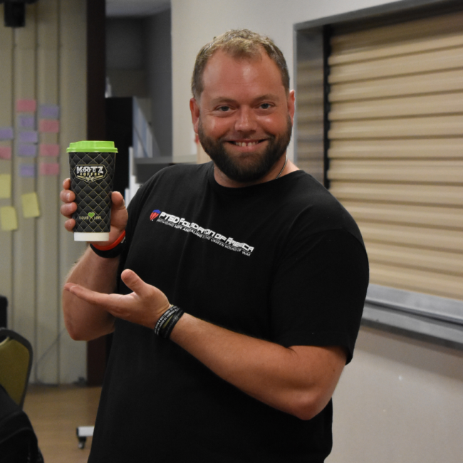 A gentleman in a PTSD Foundation of America tshirt holding a to-go cup of coffee with the Katz logo on it. He is smiling and showcasing the coffee cup.