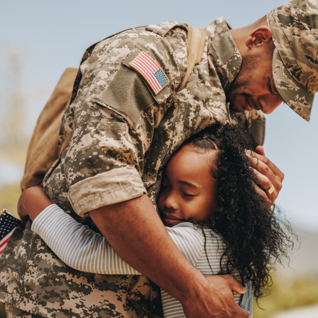 An army veteran dressed in uniform hugging his young daughter who is holding a flag.