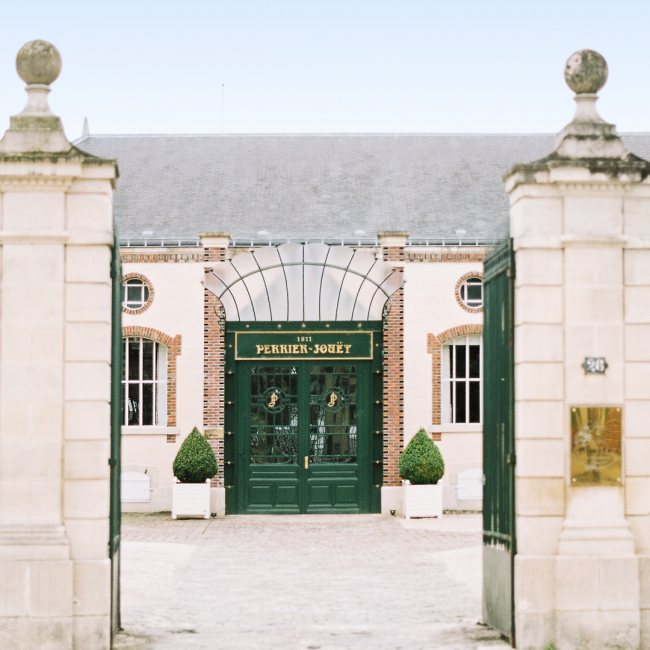 The stone entryway to the Perrier Jouet winery featuring a double-paned glass door painted in a dark green flanked by topiary plants