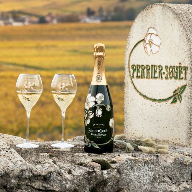 Perrie Jouet Belle Epoque Champagne bottle painted in white carnations alongside 2 glasses of champagne that have white carnations painted on them. These are sitting on a stone wall with Perrier Jouet engraved into the stone in green and white.