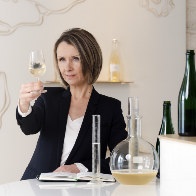 A winemaker holding up a glass of champagne studying the color alongside a beaker to measure the champagne.