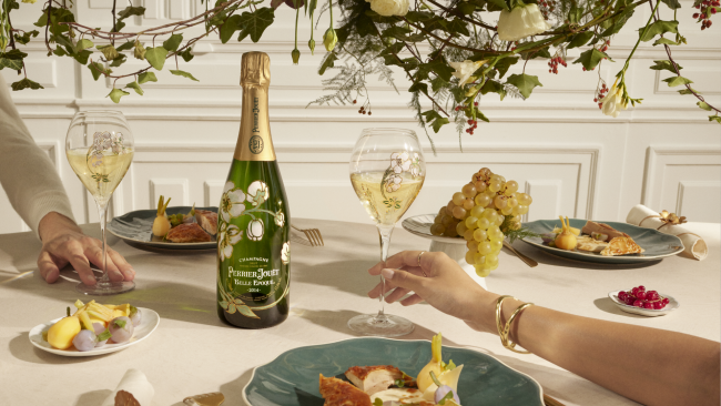 A table setting with 3 plates filled with fruits and cheese with a man and woman's hands reaching for a hand painted champagne glass filled with champagne. A bottle of Perrier Jouet Belle Epoque Champagne is in the enter with fresh florals above.