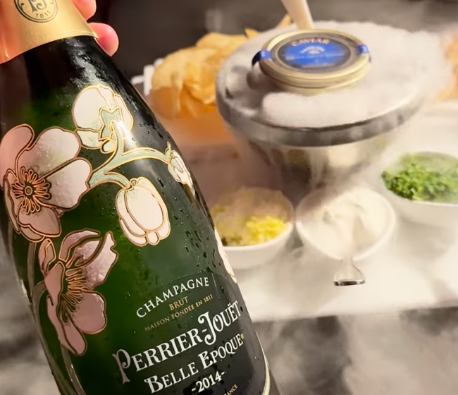 a bottle of Perrier Jouet Belle Epoque Brut Champagne vintage 2014 with TRU Caviar in the background with the traditional accoutrements and dry ice smoke.