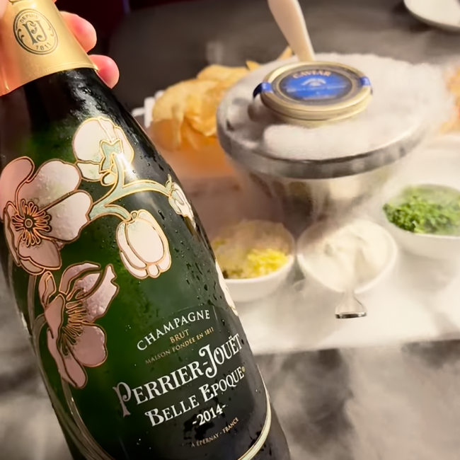 a bottle of Perrier Jouet Belle Epoque Brut Champagne vintage 2014 with TRU Caviar in the background with the traditional accoutrements and dry ice smoke.