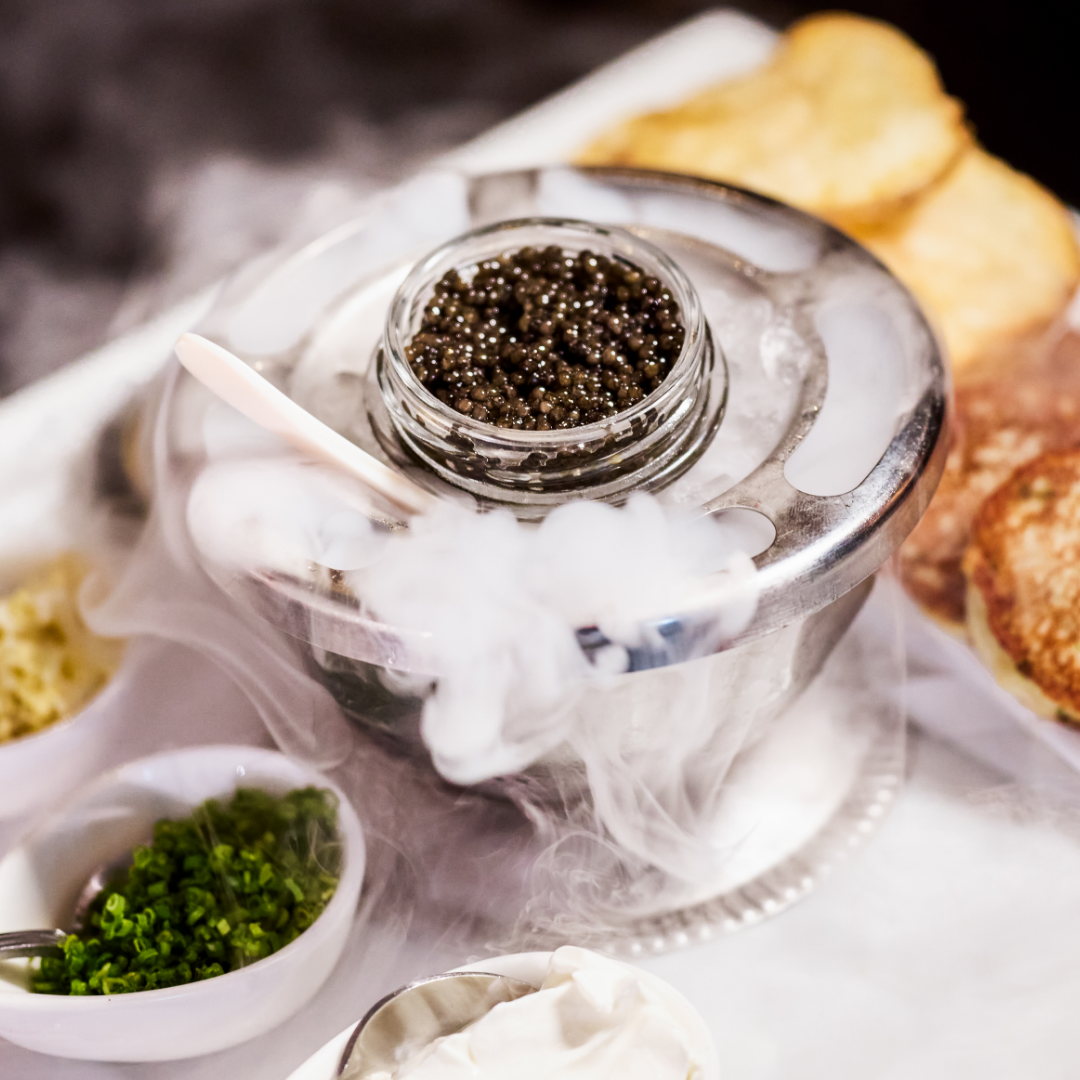 TRU Caviar in a silver container with dry ice smoke coming out of the container. There are chives and blinis flanking the caviar.