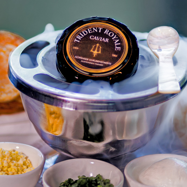 Trident Royale Caviar 28 G, acipenser gueldenstardth in a metal tin with dry ice smoke coming out of the container, a pearl spoon and chopped eggs, chives and creme fraiche in the fore ground