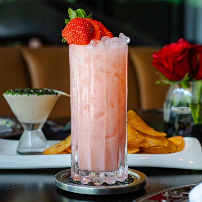 The strawberry patch a light pink non alcoholic drink in a tall glass over crushed ice with a whole strawberry sliced and fanned over top. Red roses and caviar chive dip are in the background.