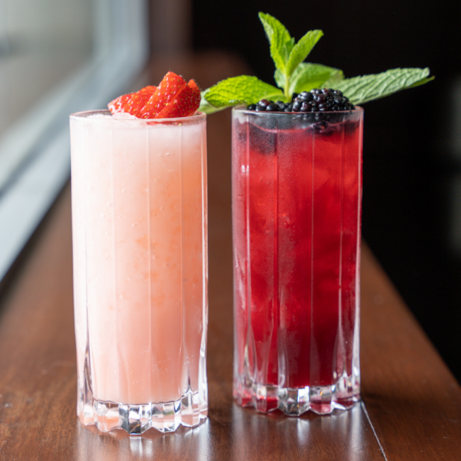 the strawberry patch zero proof cocktail in a light pink color in a tall glass topped with a strawberry fanned over top next to the blackberry bramble in a deep red color in a tall glass and topped with fresh blackberries and fresh mint.