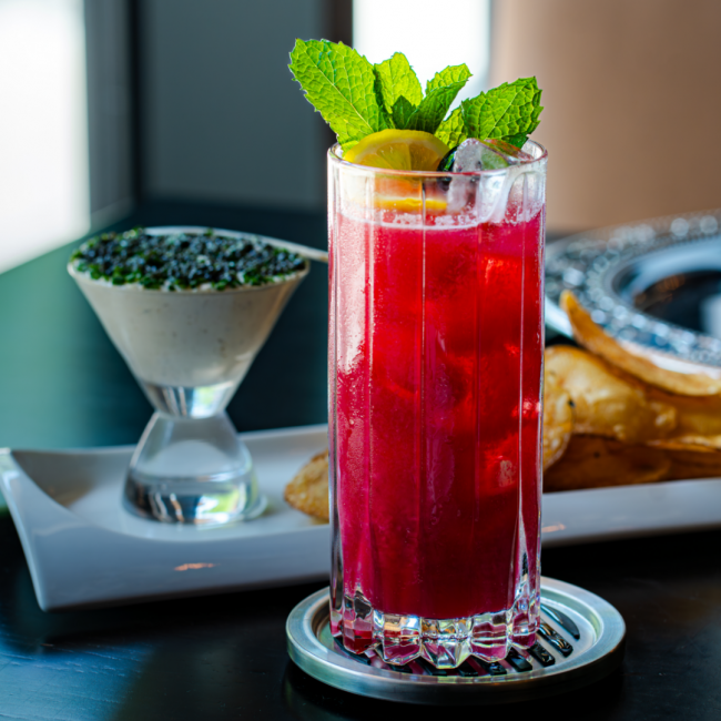 the hibiscus fresca zero proof cocktail in a tall ridged glass filled with ice cubes and topped with a lemon wheel and fresh mint. The caviar and chive dip is in the background with fresh potato chips.