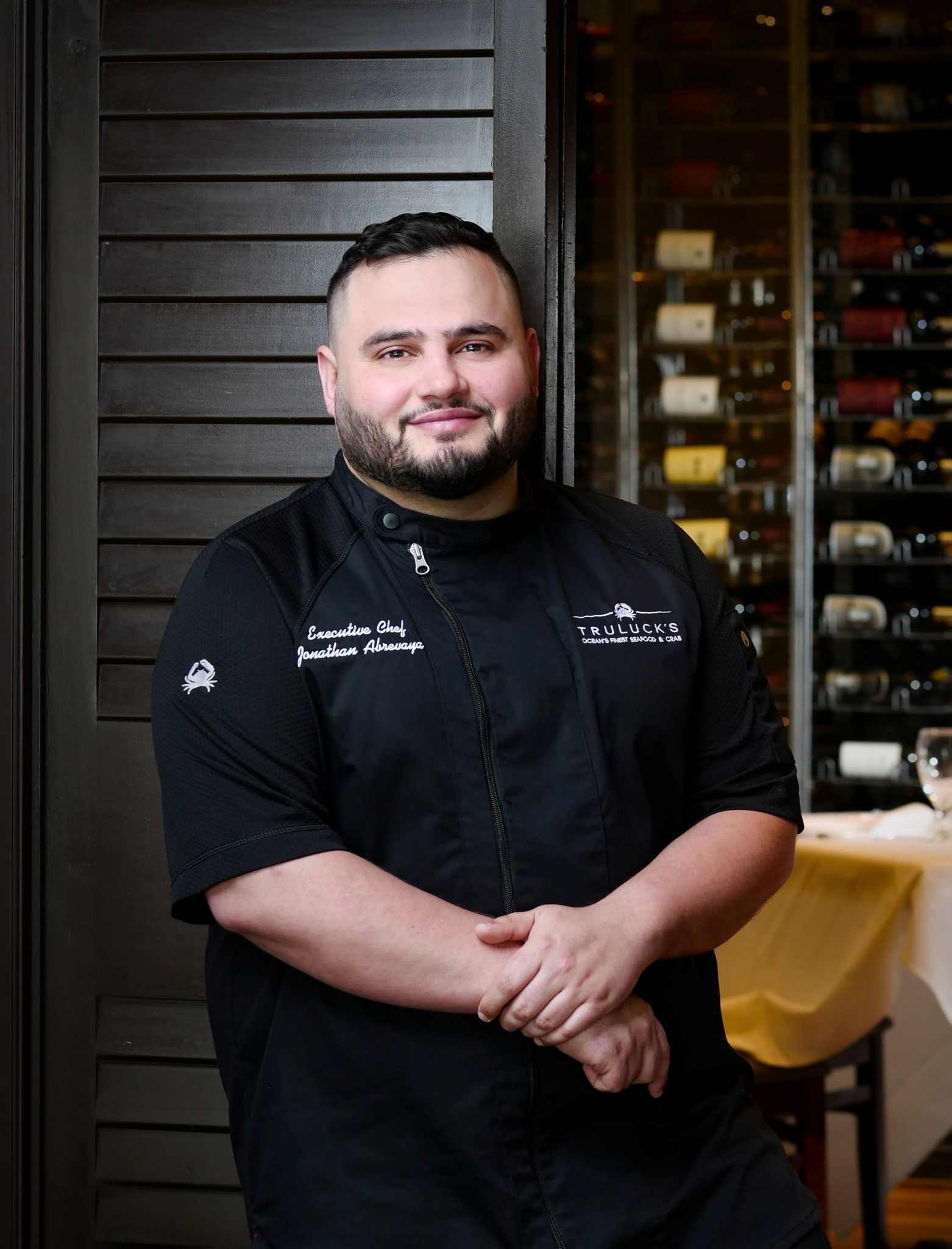 Headshot of Chef Jonathan Abrevaya in the dining room in front of a wooden shutter style door with a wine wall in the background