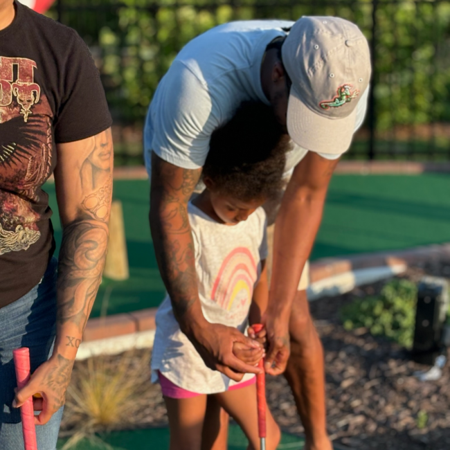 Chef Darryl showing his daughter how to hold a golf club at a mini golf place