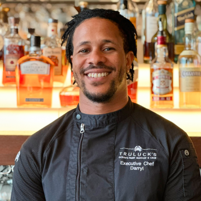 headshot of Chef Darryl Abraham in a black chef coat in front of the bar with whiskey bottles in the background