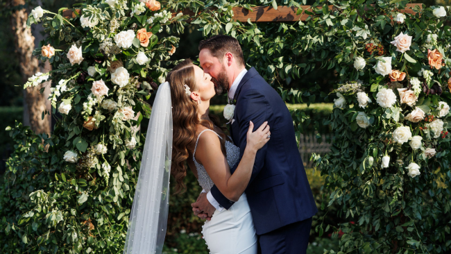 Karen and Chris in wedding attire sharing a first kiss under a terrace of greenery filled with white and peach roses.