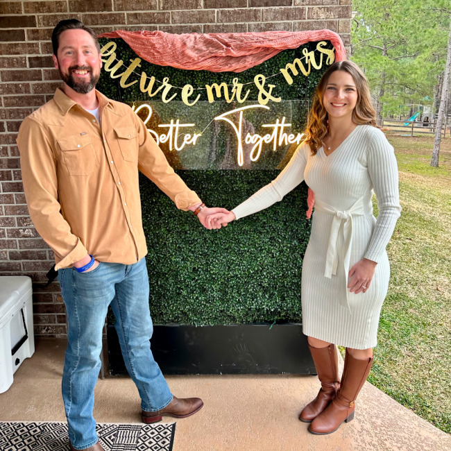 Chris and Karen standing in front of a photo backdrop that says "future mr & mrs, better together" at a backyard engagement party.