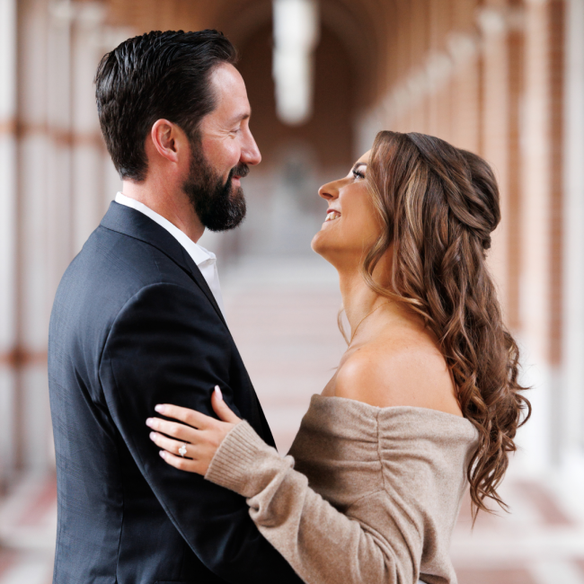 Chris and Karen's engagement photo with them facing each other and smiling. Chris is wearing a dark suit and Karen is wearing an off the shoulder tan sweater dress