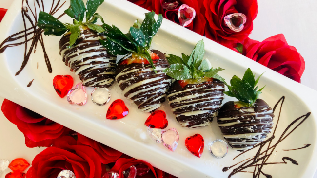 Four white and dark chocolate covered strawberries on a white rectangular plate with red roses in the background