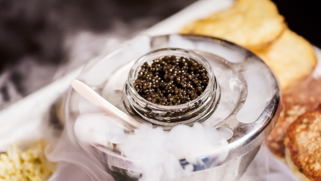 A close up shot of a jar of fresh caviar with dry ice smoke coming out of the vessel full of ice with a pearl spoon in the ice. Warm blinis and potato chips are in the background.