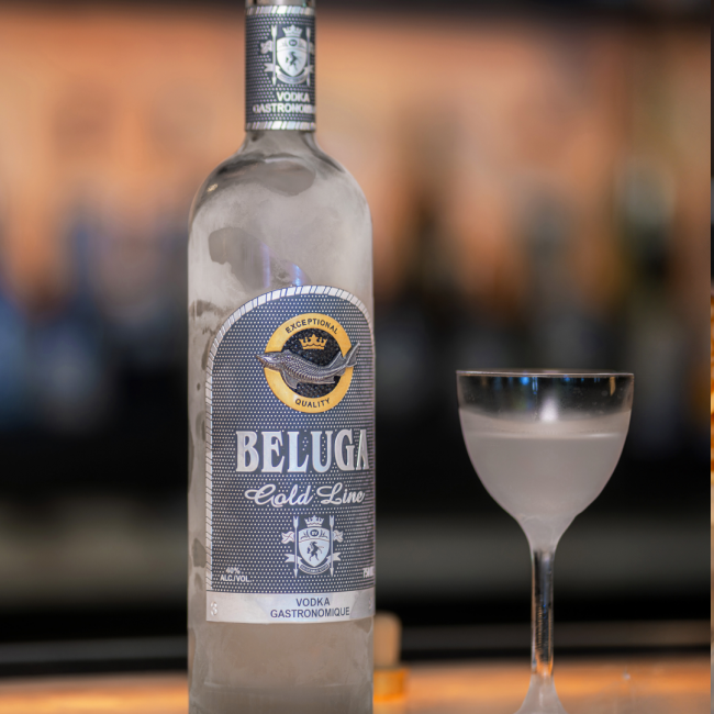 An ice cold bottle of Beluga Gold Line vodka on the bar top next to a chilled Nick & Nora style glass filled with icy cold vodka.