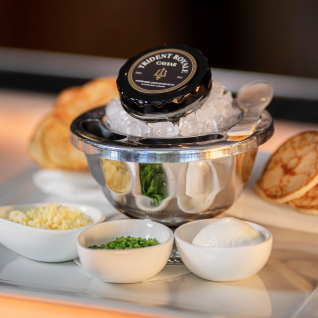 Trident Royale Caviar tin on ice in a silver vessel with a pearl spoon surrounded by small dishes of grated egg, chopped chives, creme fraiche and warm blinis.