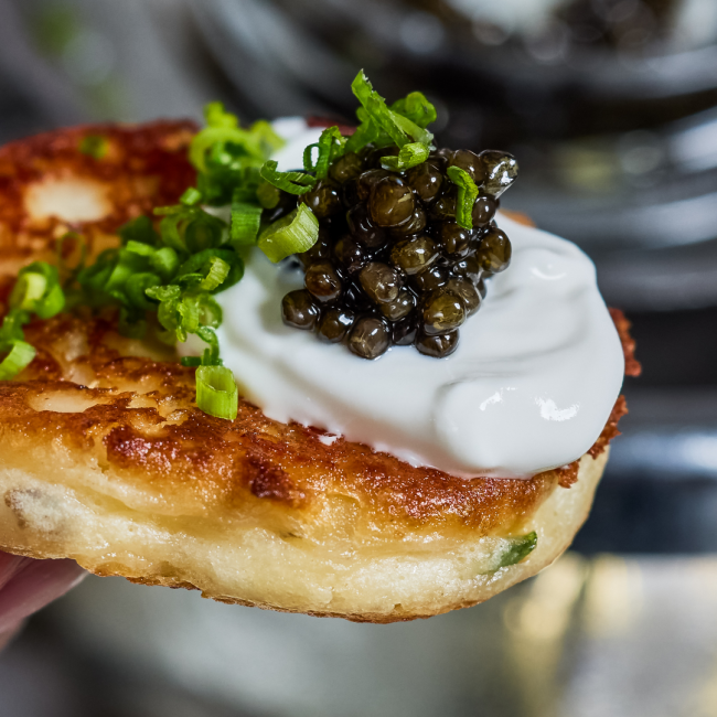 A close up image of a a warm blini topped with creme fraiche, TRU Caviar and chives.