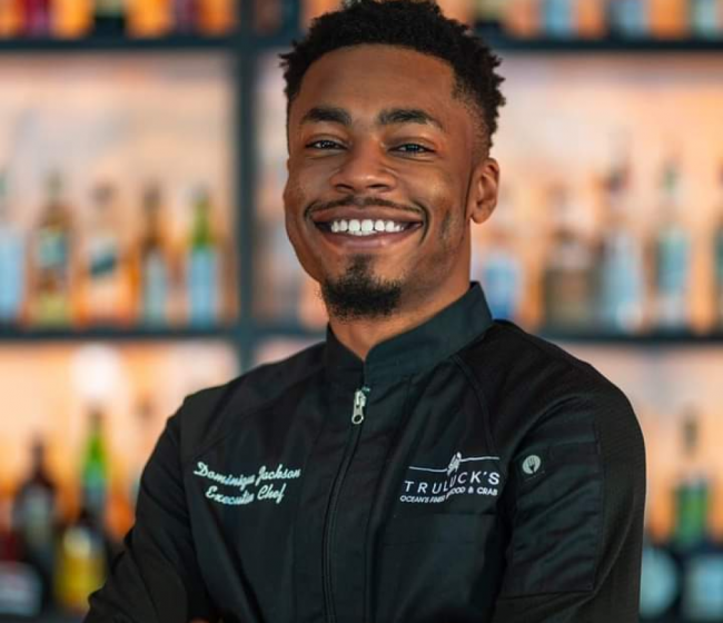 Headshot of Chef Dom in his black chef coat standing in front of the bar top smiling