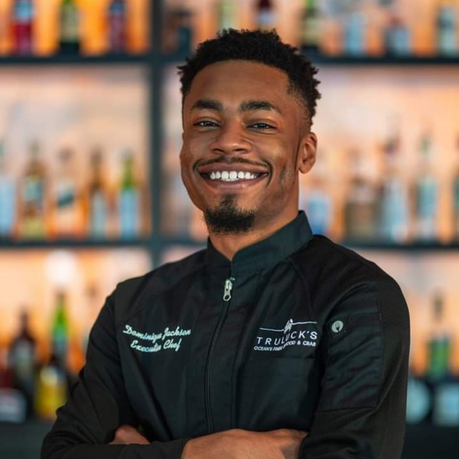 Headshot of Chef Dom in his black chef coat standing in front of the bar top smiling