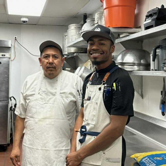 Chef Dom and Cam in the Arboretum kitchen smiling and wearing aprons