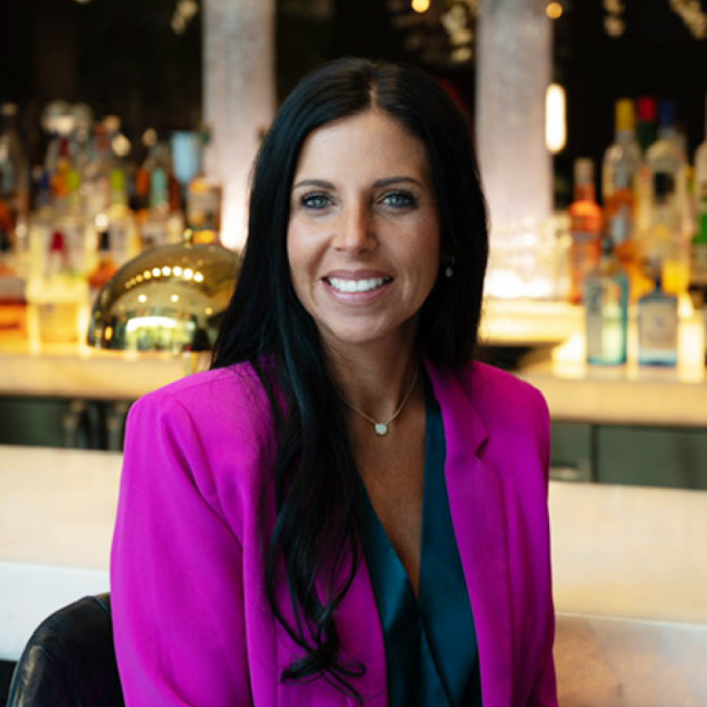 headshot of general manager Tambra Tatum in a blue blouse with bring pink blazer smiling and sitting in front of the bar top