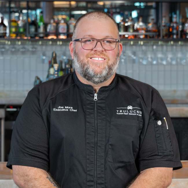 headshot of Chef Joe Mora in a black chef coat with a white Truluck's logo in front of the bar