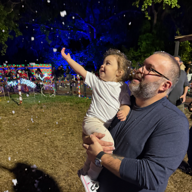 Chef Joe Mora holding his little girl up at a Christmas event while she catches faux snow in the air