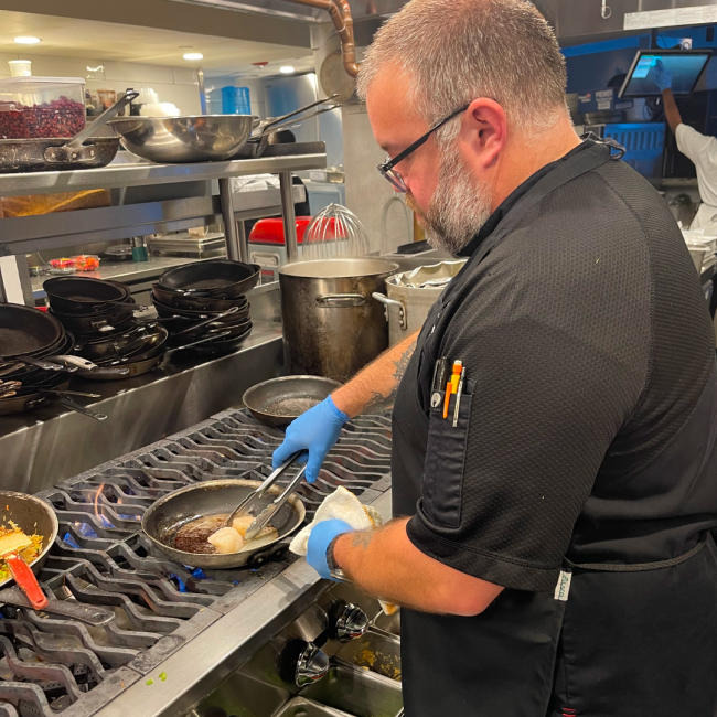 Chef Joe Mora in a black chef coat on the line cooking scallops in a sautee pan