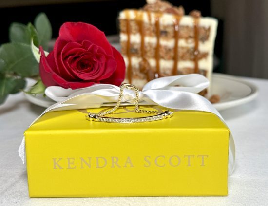 Kendra Scott yellow gift box with a gold Ott bracelet on top of the box with a red rose and a large slice of carrot cake in the background