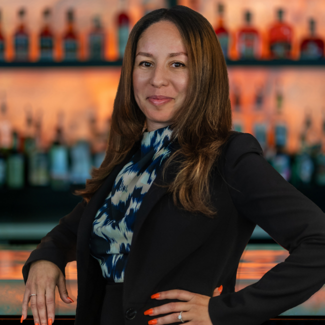 Myriam Chiny Truluck's Arboretum general manager in a dark suit with a floral top standing in front of the bar top with her elbow on the bartop and hand on her hip.