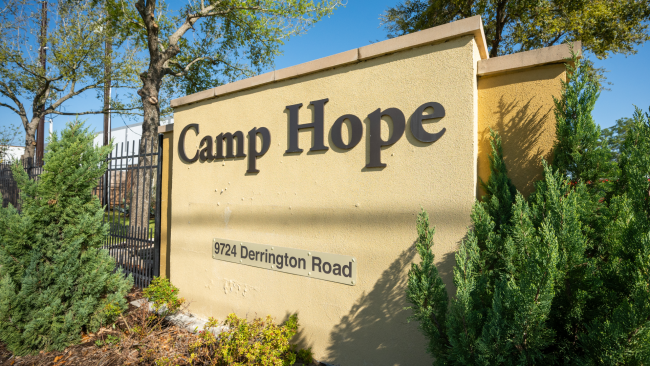 The entryway sign that says "Camp Hope 9724 Derrington Road" flanked by two juniper bushes