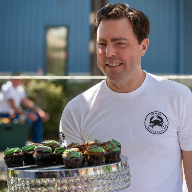 Dave Mattern in a Camp Hope tshirt carrying a riser of cupcakes with camo frosting