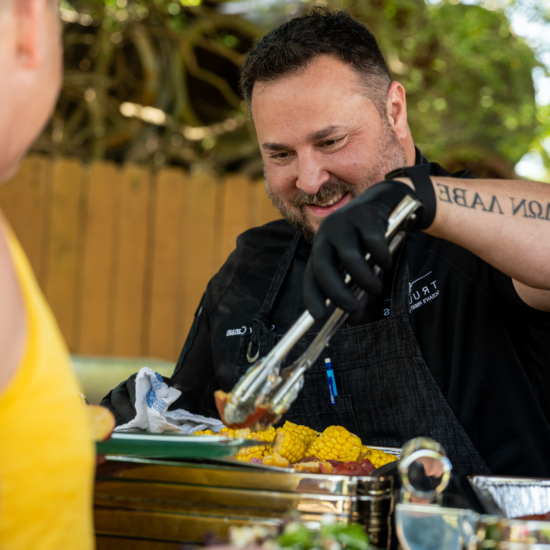 Chef Sammy in a black chef coat smiling and serving roasted corn and potatoes with tongs to a guest from a silver chafing dish