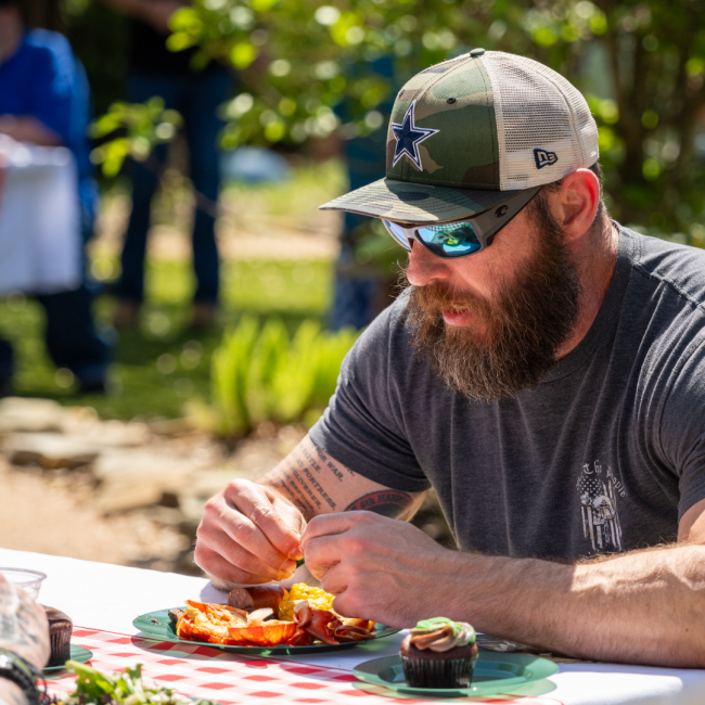 a veteran resident of Camp Hope enjoying a lobster boil lunch with a chocolate cupcake at a picnic table outside dressed in a Dallas Cowboys camo hat and sunglasses