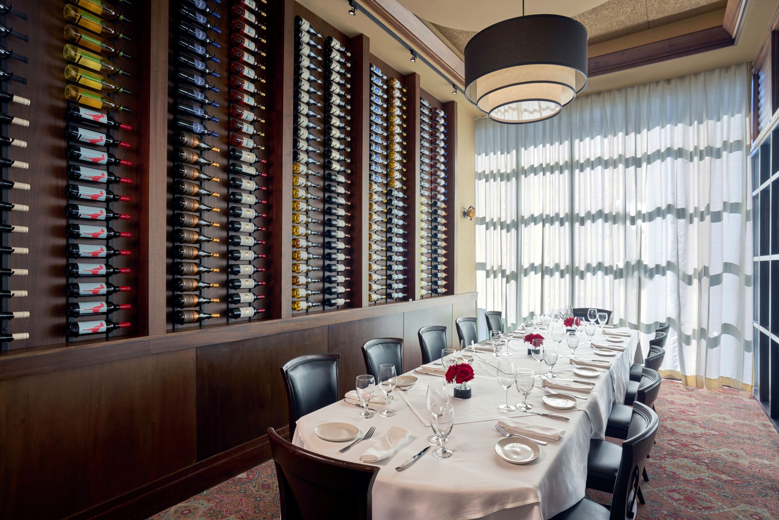 The Big Deal Room - an elegant private dining room with a long white-tablecloth table set for twelve, featuring dark wood chairs, a wall-mounted wine display with colorful bottles, a large circular light fixture, and floor-to-ceiling sheer curtains filtering natural light.