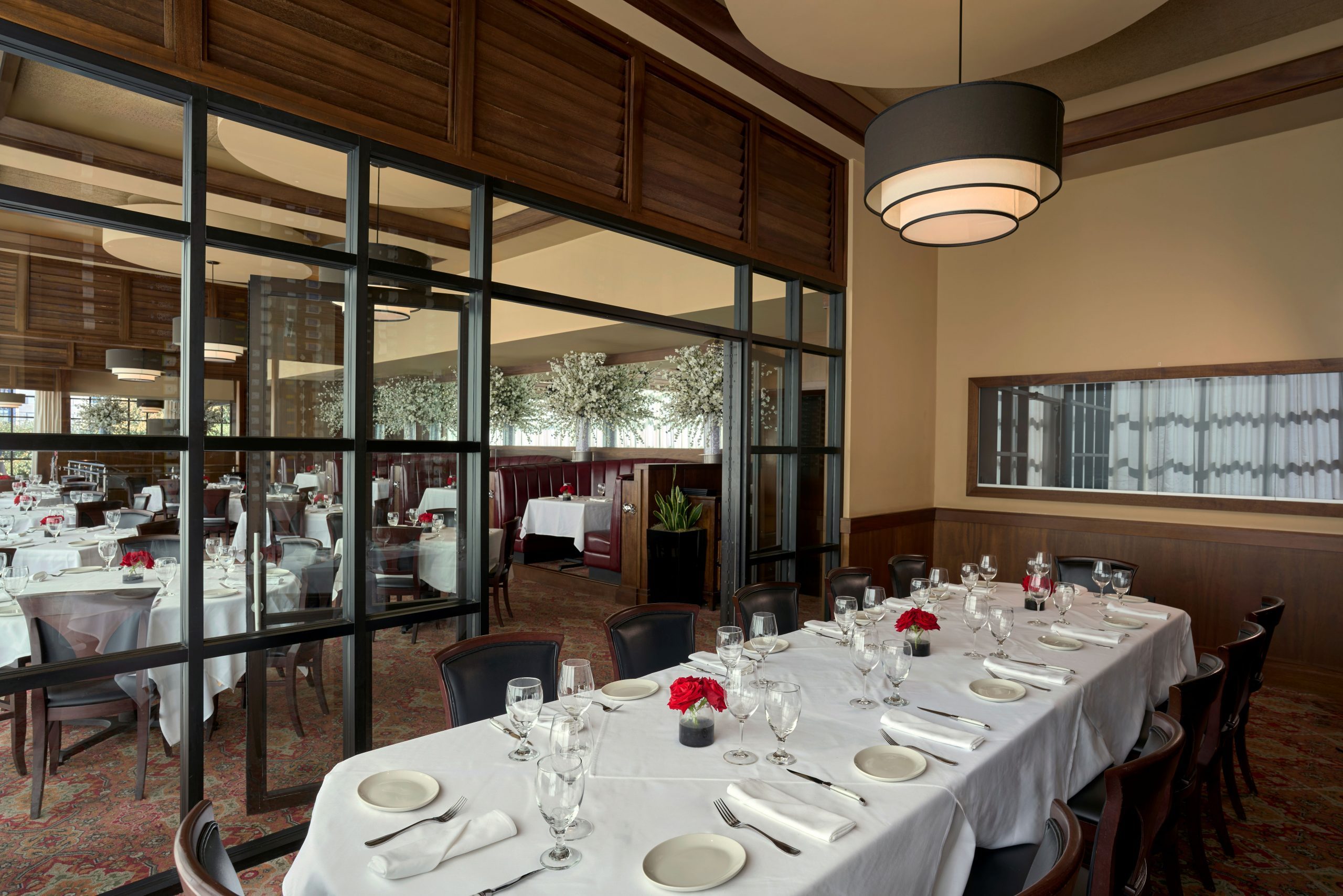The Big Deal Room - a refined private dining room with a long table set for twelve, dressed in a white tablecloth and accented with red rose centerpieces. The space features dark wood paneling, a modern tiered chandelier, and glass-paneled walls that offer a view into the elegant main dining area with booth seating and floral arrangements.
